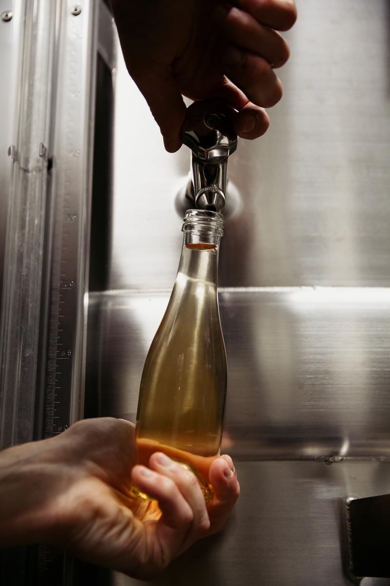 Close up of hands filling a sample bottle of wine from a stainless steel vat